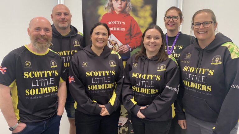 A group of six people smiling, wearing "Scotty's Little Soldiers" hoodies, stands in front of a banner featuring a child in a red hoodie. Their presence reflects Scotty’s commitment to supporting bereaved adults and children alike.