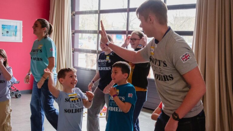 A group of children and adults wearing "Scott's Little Soldiers" shirts, part of charities supporting bereaved families, interact and high-five in a bright indoor room during one of their events.
