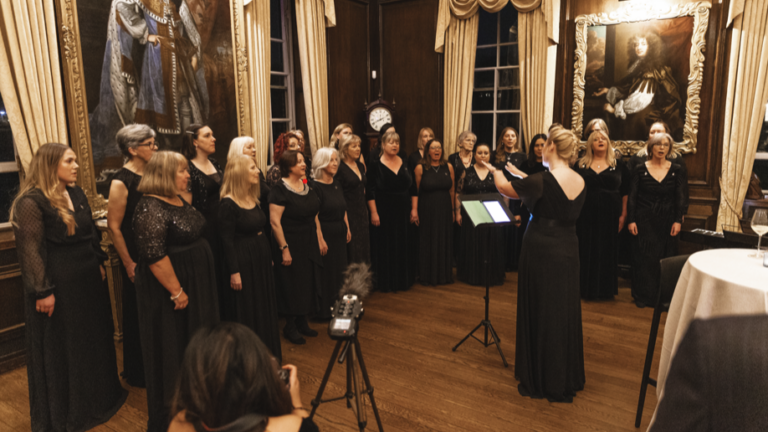 A choir of women in black dresses sings in a wood-paneled room adorned with paintings and a clock. At this Veterans’ Foundation gala, a woman conducts passionately as the microphone captures their harmonious tribute.