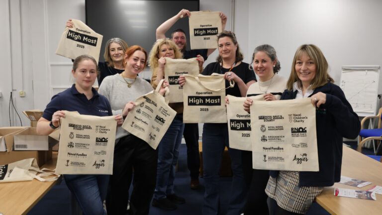A group of families holding tote bags labeled "Operation High Mast 2023" with various logos are standing in a room with a table and chairs, highlighting their commitment to collective wellbeing.
