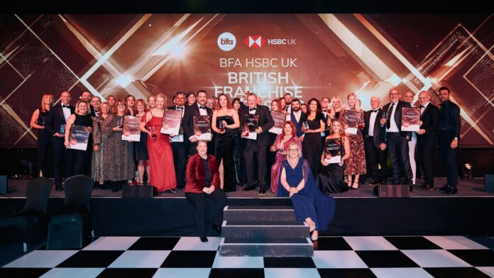 A large group of people in formal attire pose on stage holding certificates at the BFA HSBC UK British Franchise Awards event, celebrating the best in franchising.