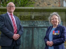 A man in a suit and a woman with medals on her blazer stand in front of the Scottish Veterans’ Memorial, a stone wall engraved with text, symbolizing efforts to restore Scottish veterans' recognition.