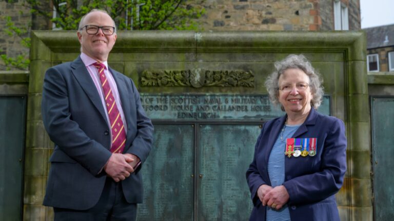 A man in a suit and a woman with medals on her blazer stand in front of the Scottish Veterans’ Memorial, a stone wall engraved with text, symbolizing efforts to restore Scottish veterans' recognition.