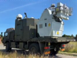 A military truck carries a large mounted system for drone defense, equipped with radar and a visible laser warning label, parked on a grassy area under a clear sky.