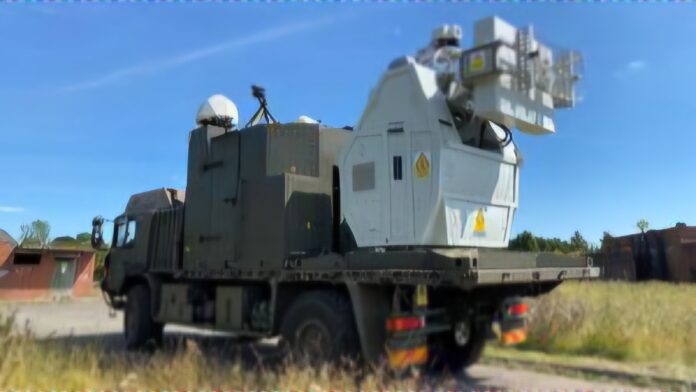 A military truck carries a large mounted system for drone defense, equipped with radar and a visible laser warning label, parked on a grassy area under a clear sky.