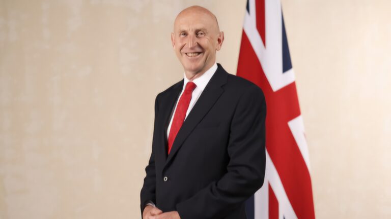 A man in a suit with a red tie stands smiling in front of a UK flag, embodying the spirit of partnership between the UK's Defence sector and the private sector.