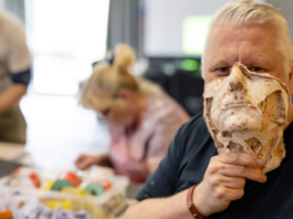 A man holds a lifelike sculpted mask to his face—an example of how Blesma empowers members through creative storytelling, while others work on crafts at a table in the background.