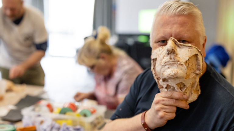 A man holds a lifelike sculpted mask to his face—an example of how Blesma empowers members through creative storytelling, while others work on crafts at a table in the background.