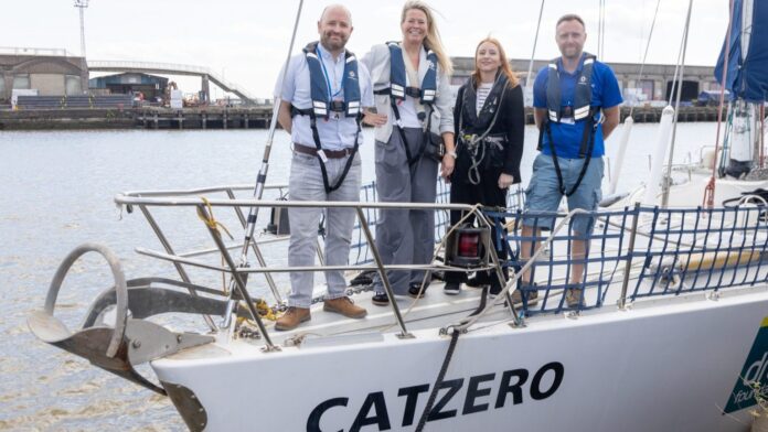 Four people wearing life vests stand on the deck of CATZERO, docked at a harbor with industrial buildings—a scene from the Family Sailing Programme, offering a life-changing experience on the water.