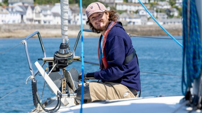 A limbless veteran wearing a cap, gloves, and a life jacket sits at the bow of a sailboat on calm water near a coastal town, taking part in the Pacific Sailing Challenge.