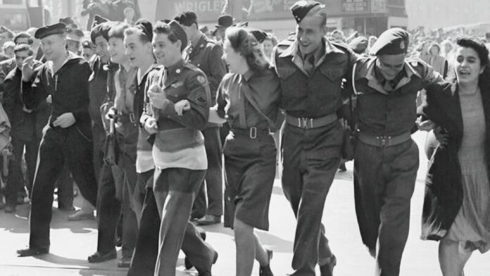A group of uniformed men and women, including Ahmadiyya Muslims, walk arm in arm down a city street, smiling and celebrating among a crowd on VE Day.