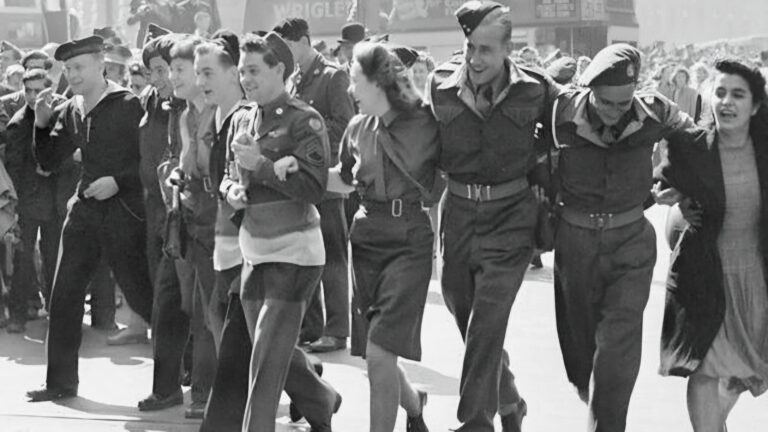A group of uniformed men and women, including Ahmadiyya Muslims, walk arm in arm down a city street, smiling and celebrating among a crowd on VE Day.