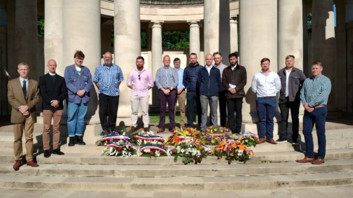 A group of 15 men stands in front of columns behind several floral wreaths and bouquets arranged on stone steps, in bright sunlight, paying tribute to Windsor Soldiers with Military Honours.