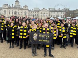 A large group of military children and teens, many wearing black and yellow scarves, stand together holding a "Scotty's Little Soldiers" banner in an outdoor urban setting.