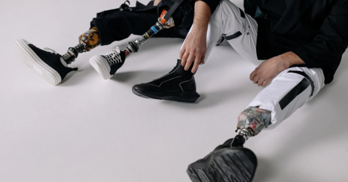 Two people with limb loss sit on the floor, wearing black and white sneakers and casual pants, their prosthetic limbs clearly visible.