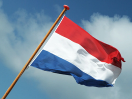 A red, white, and blue horizontal tricolor flag on a wooden pole flying against a partly cloudy blue sky, representing the spirit of Taxi Charity and celebrating its recent Royal Honour.