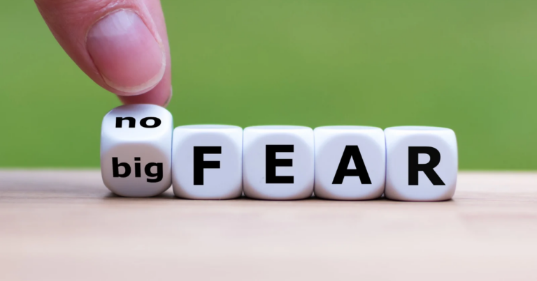 A hand flips a cube to change the phrase from "big FEAR" to "no FEAR" on white blocks with black letters, promoting the Free from Fear Programme against a wooden surface and green background.