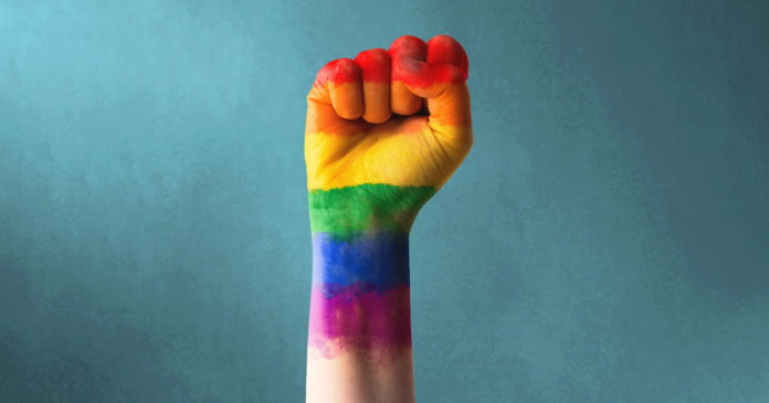 A raised fist with a rainbow flag painted on the forearm against a blue background, symbolizing pride and resilience—an image resonating with tributes following the obituary of Lord Etherton.