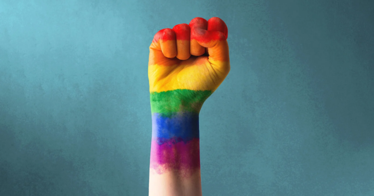A raised fist with a rainbow flag painted on the forearm against a blue background, symbolizing pride and resilience—an image resonating with tributes following the obituary of Lord Etherton.