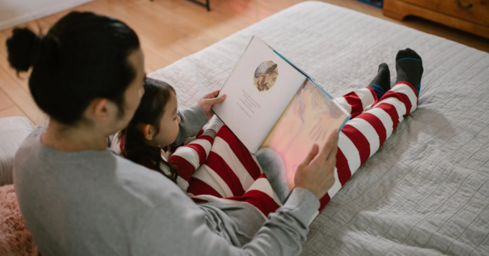 An adult and a child, both in striped pajamas, sit on a bed reading a picture book together, sharing the joy of kids reading before bedtime.