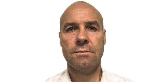 A bald man with fair skin and blue eyes, wearing a white shirt, faces the camera against a plain white background, reflecting a professional look suited to project management or an Army career.