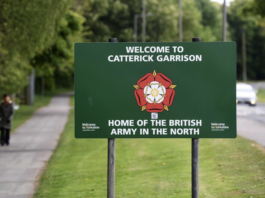 Green sign reads "Welcome to Catterick Garrison, Home of the British Army in the North" with a red and white rose emblem. The Catterick landmark stands beside a road lined with trees in the background.