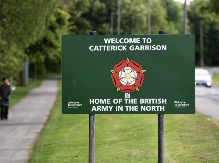 Green sign reads "Welcome to Catterick Garrison, Home of the British Army in the North" with a red and white rose emblem. The Catterick landmark stands beside a road lined with trees in the background.