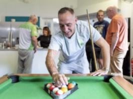 A man arranges billiard balls on a pool table while four other people talk in the background in a kitchen area.