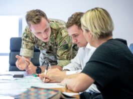 A person in military uniform discusses documents with two people in civilian clothing at a table, highlighting the collaboration between employers and the Armed Forces community in an office setting.