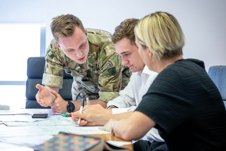 A person in military uniform discusses documents with two people in civilian clothing at a table, highlighting the collaboration between employers and the Armed Forces community in an office setting.