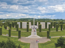 A circular outdoor memorial honoring LGBT+ Armed Forces stands among tall trimmed trees and stone walls, with a central obelisk. Community voices gather here, surrounded by green lawns under a partly cloudy sky.