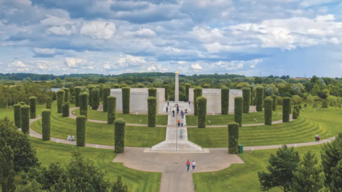 A circular outdoor memorial honoring LGBT+ Armed Forces stands among tall trimmed trees and stone walls, with a central obelisk. Community voices gather here, surrounded by green lawns under a partly cloudy sky.
