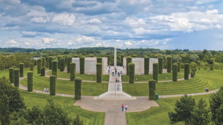 A circular outdoor memorial honoring LGBT+ Armed Forces stands among tall trimmed trees and stone walls, with a central obelisk. Community voices gather here, surrounded by green lawns under a partly cloudy sky.