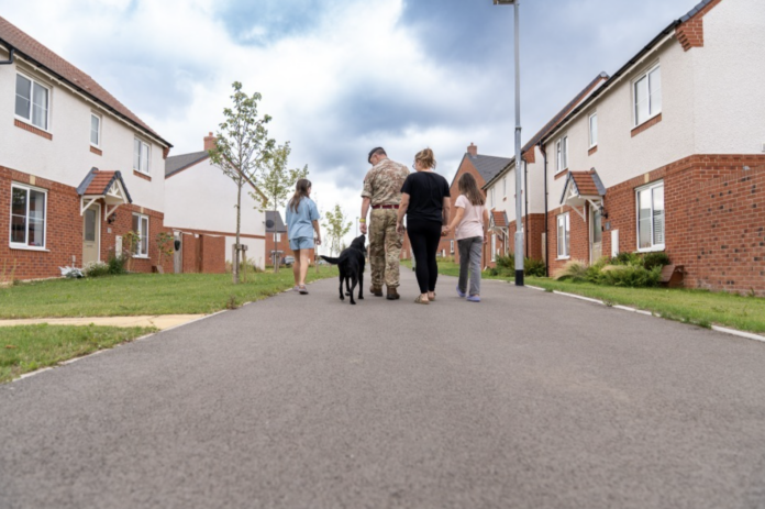 A man in military uniform walks with three women and a black dog down a residential street lined with detached houses, reflecting the community spirit fostered by Armed Forces charities like the Veterans’ Foundation.