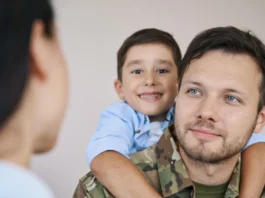 A man in military uniform with a young boy smiling on his back faces a woman in the foreground, highlighting veterans and their transition to civilian employment.