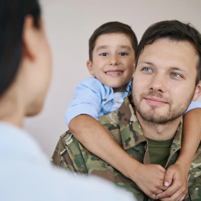 A man in military uniform with a young boy smiling on his back faces a woman in the foreground, highlighting veterans and their transition to civilian employment.
