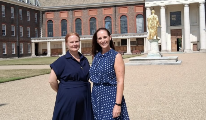 Two women in blue dresses stand outside a historic building with columns and a gold statue, honoring discharged UK Armed Forces members on a sunny day.