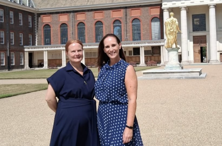 Two women in blue dresses stand outside a historic building with columns and a gold statue, honoring discharged UK Armed Forces members on a sunny day.