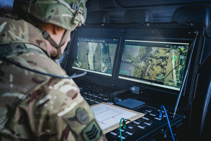 A soldier in camouflage uniform monitors aerial surveillance footage from AI-powered drones on dual screens inside a military vehicle, working to detect landmines and other explosive threats.