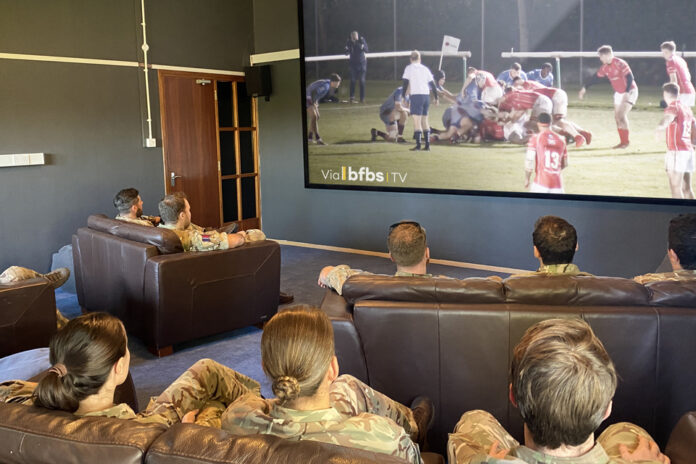 Several people in military uniforms, gathered for veterans' wellbeing, sit on sofas watching a rugby match on a large screen in a room.