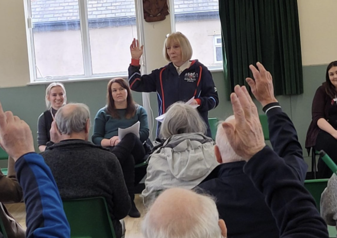 A woman stands with her hand raised, addressing a seated group of people, some of whom also have their hands raised, in a community hall.