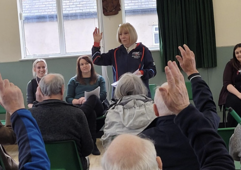 A woman stands with her hand raised, addressing a seated group of people, some of whom also have their hands raised, in a community hall.