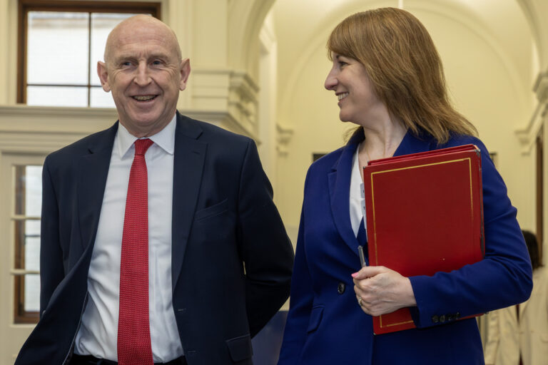A man in a suit and red tie stands next to a woman in a blue suit holding a large red folder as they walk and talk in a building with arched doorways, discussing how to explain military experience for better civilian understanding.