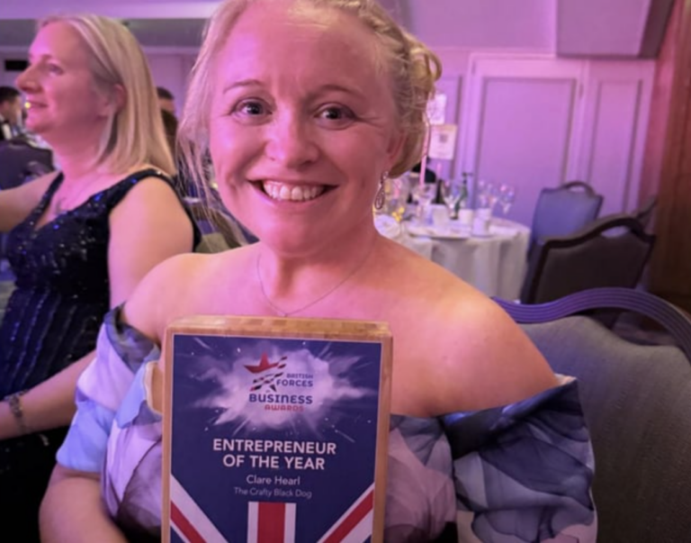 A woman in a blue floral dress smiles and holds a plaque reading "Entrepreneur of the Year" at an indoor awards event, honoured for her craft business with tables visible in the background.
