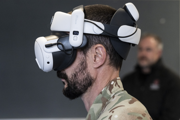 A man in military camouflage wears two stacked virtual reality headsets, viewed from the side, with another person blurred in the background.