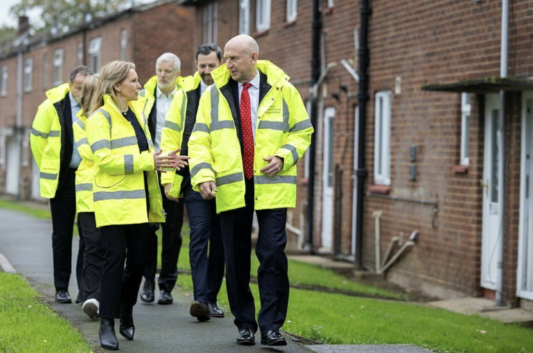 A group of people wearing high-visibility jackets walk and talk along a pavement in front of a row of brick military homes.