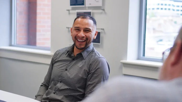 Peter Alford, wearing a patterned shirt, sits at a table smiling in a bright office with large windows and wall-mounted plaques, reflecting on his career change from construction.