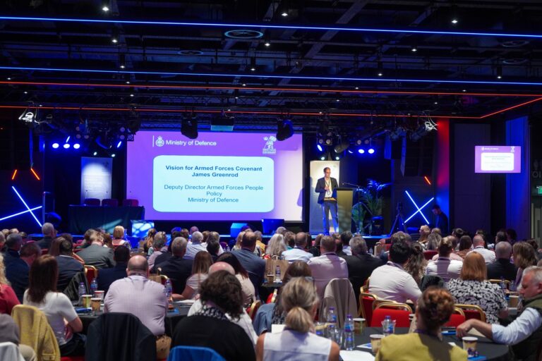 A speaker presents on stage to a seated audience at a Ministry of Defence Conference 2026 event, with a presentation slide titled "Vision for Armed Forces Covenant" displayed behind him.