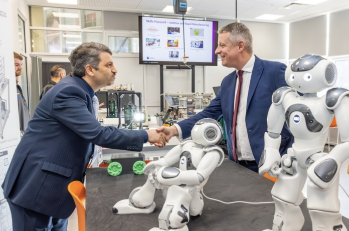 Two men in suits shake hands in a tech lab, with two humanoid robots and equipment on the table—highlighting how universities are advancing defence skills through cutting-edge research and innovation.