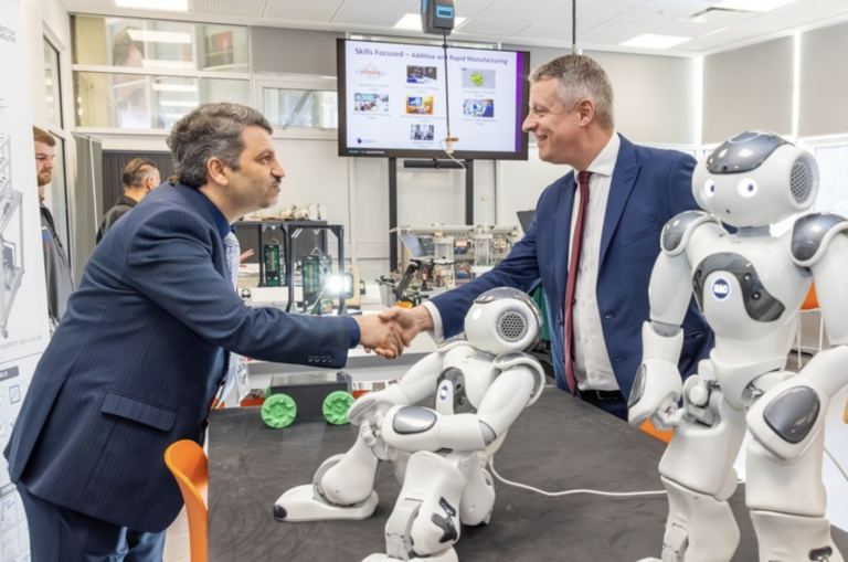 Two men in suits shake hands in a tech lab, with two humanoid robots and equipment on the table—highlighting how universities are advancing defence skills through cutting-edge research and innovation.